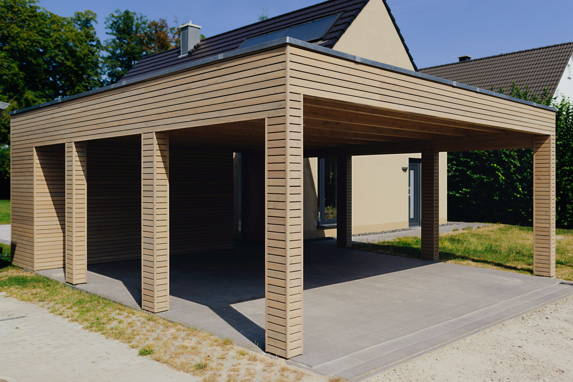Ein großer Carport aus Holz befindet sich vor einem gelbgestrichenen Einfamilienhaus.