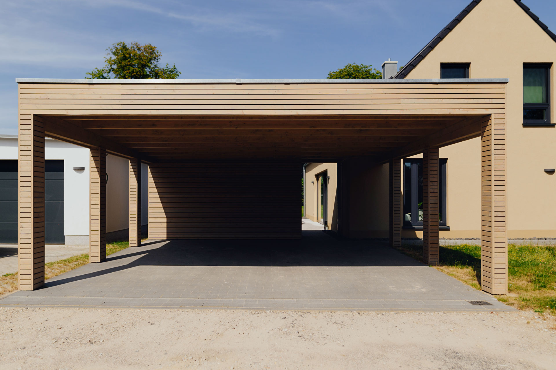 Ein großer Carport aus Holz befindet sich vor einem gelbgestrichenen Einfamilienhaus. Die Sonne scheint.