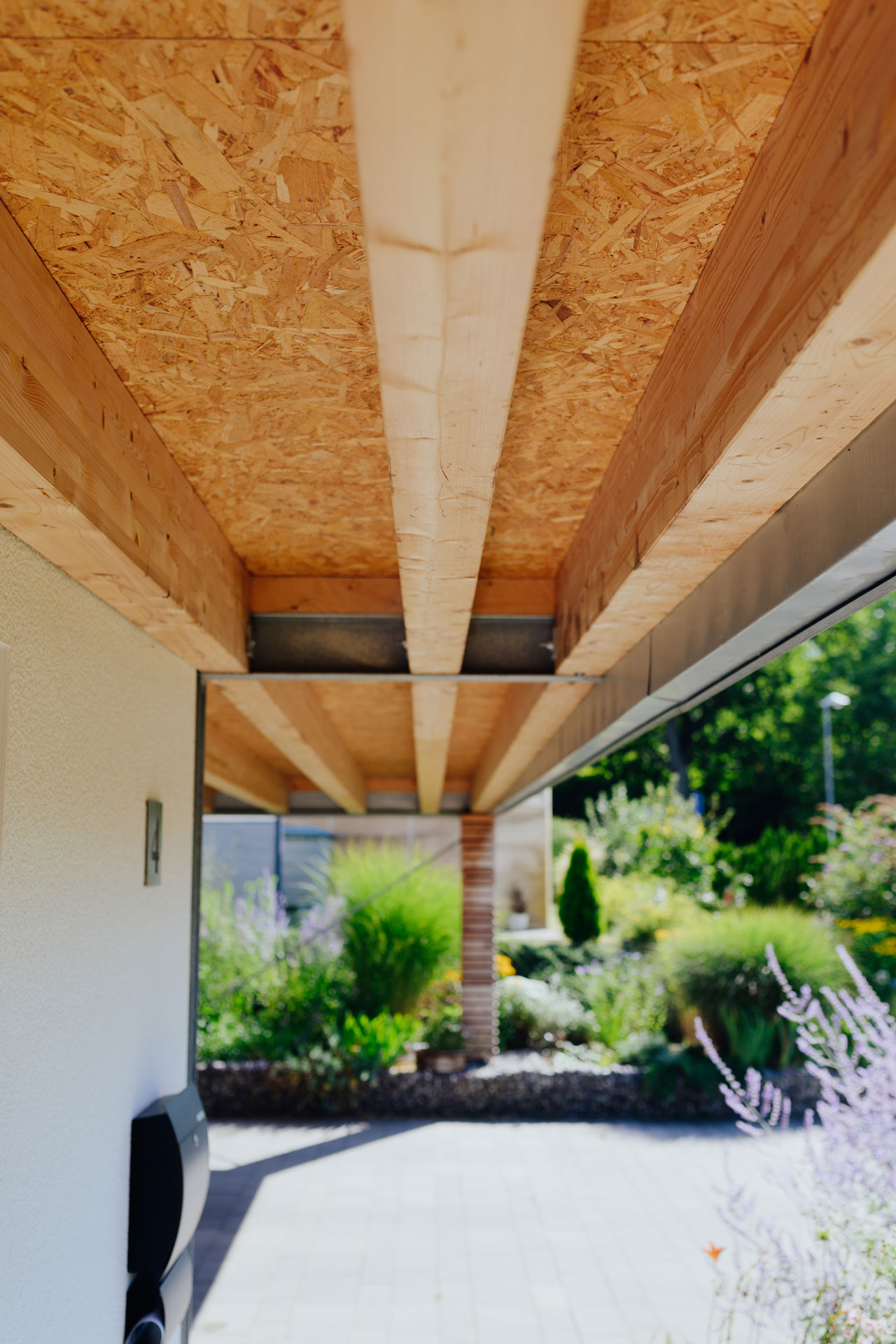 Massive Holzbalken stützen ein Carportdach. Die Sonne bescheint den nebenstehenden Lavendel.