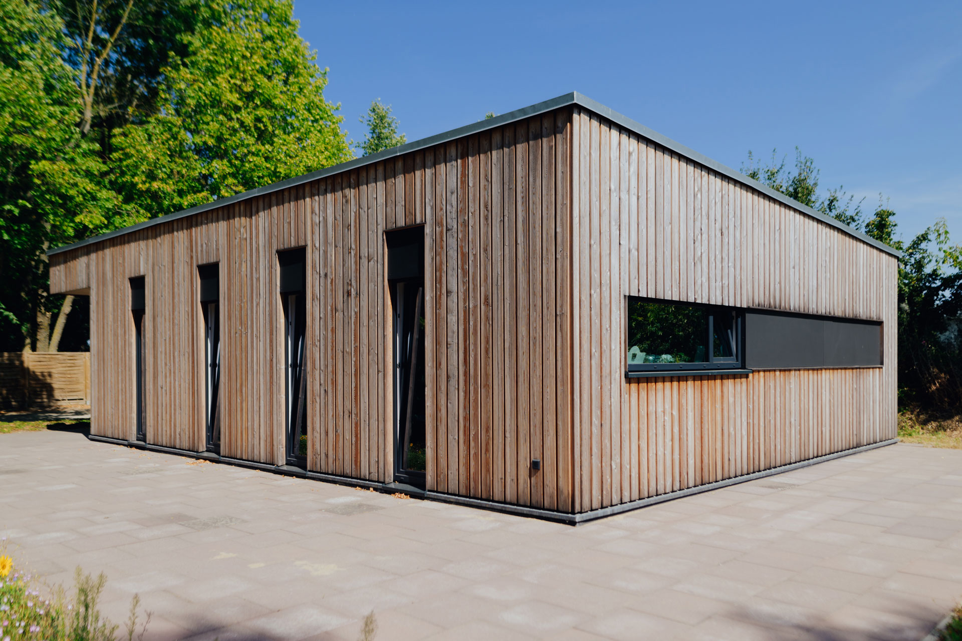 Eine Schuppenanlage aus Holz hat auf der einen Seite vier vertikale und auf der anliegenden Seite ein horizontal schmales Fenster.