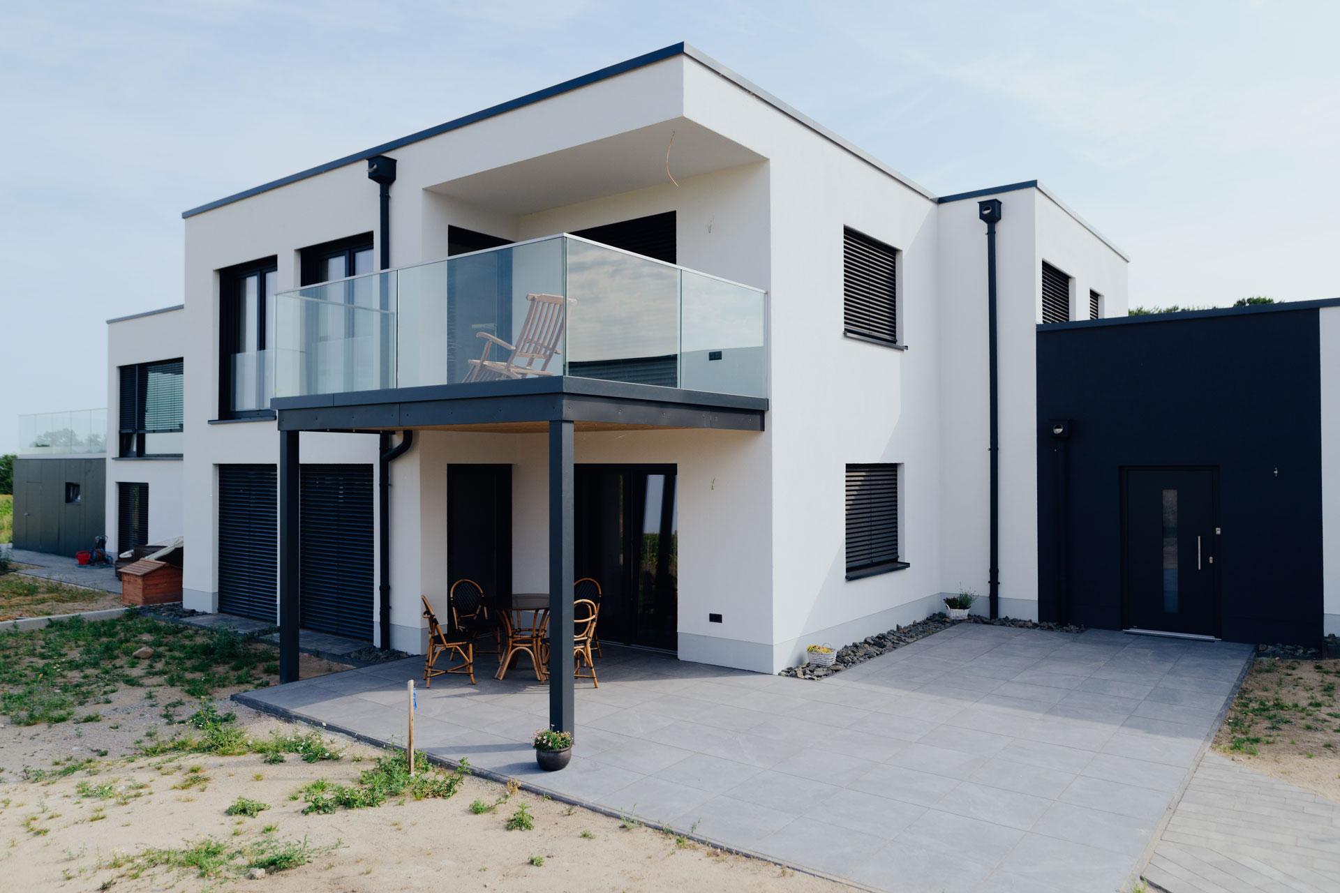 Auf einer Terrasse eines modernen Doppelhauses steht ein Tisch und fünf Stühle. Im Hintergrund ist ein Feld zu erahnen.
