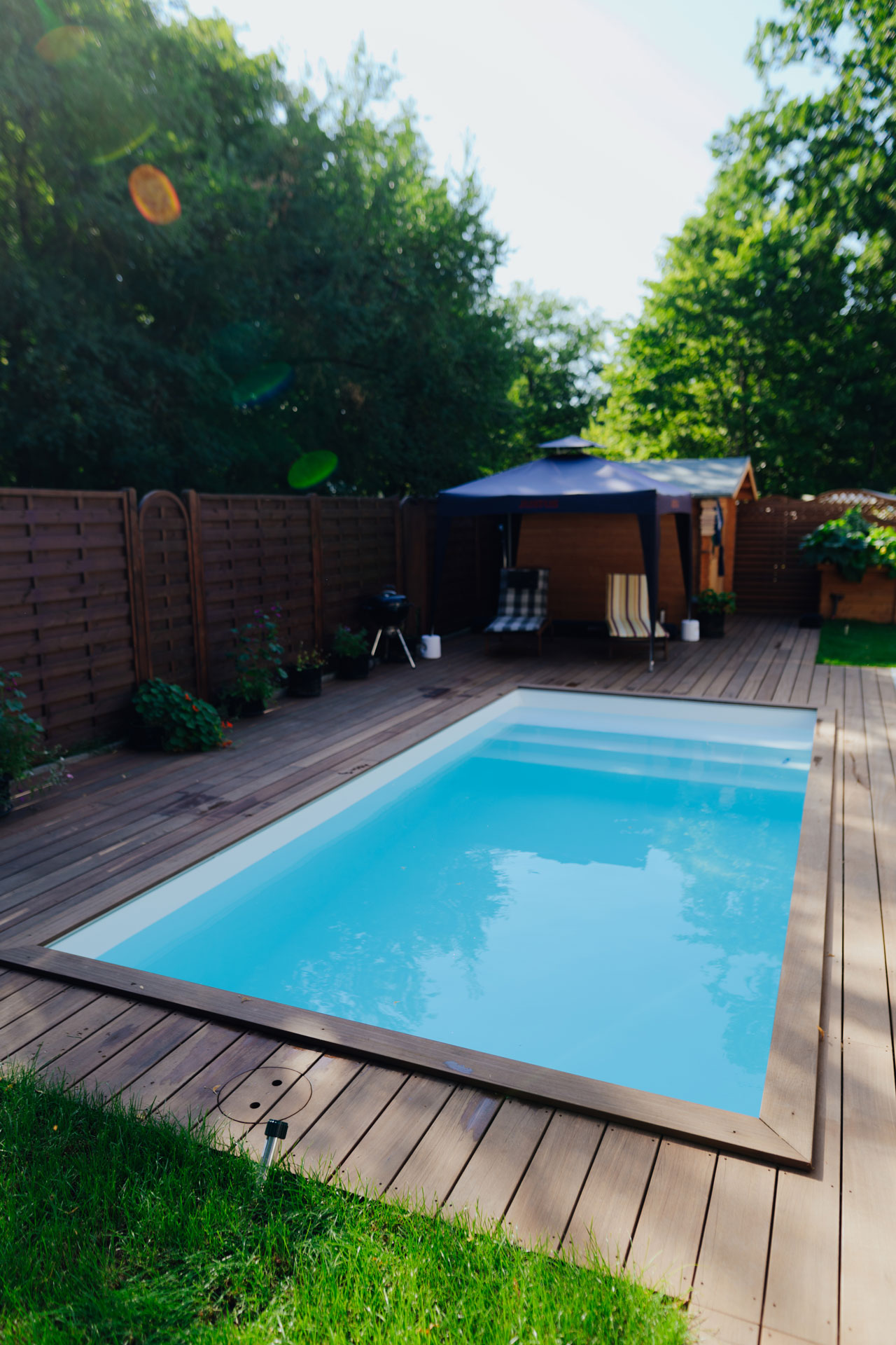 Eine Terrasse aus Holz hat einen rechteckigen Pool. Im Hintergrund steht ein kleines Gartenhaus.