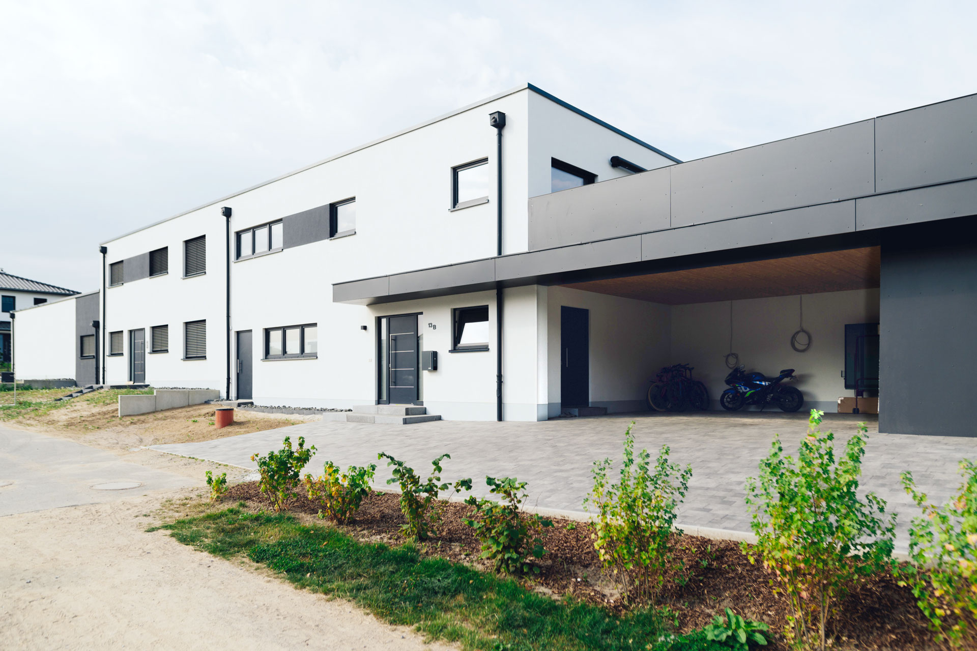 Ein Doppelhaus hat einen großen angrenzenden Carportanbau. Eine Terrasse befindet auf dem anthrazitfarbenen Garagenanbau.