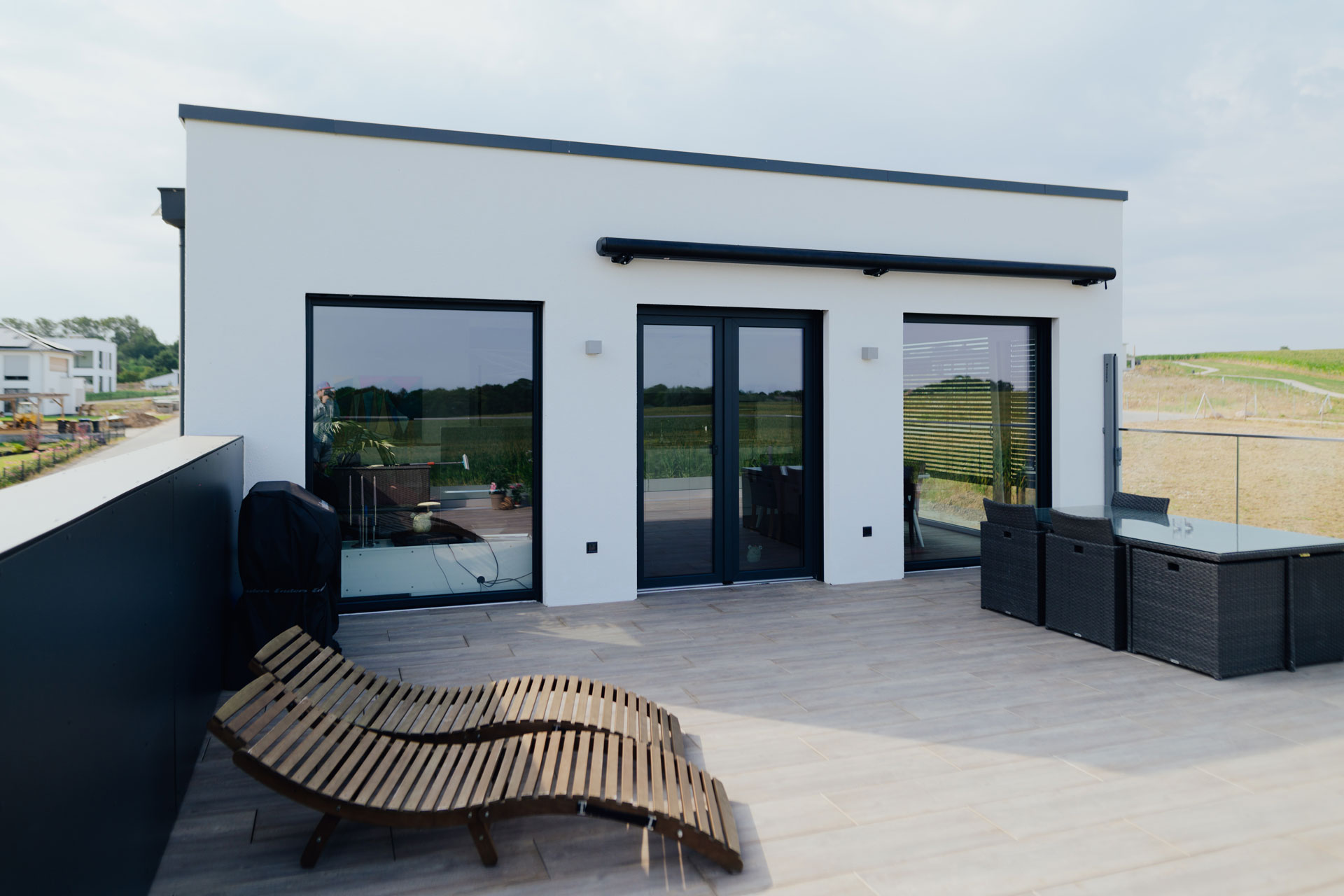 Auf einer Dachterrasse sind zwei Sonnenliegen aus Holz. In den Fenstern des Gebäudes spiegelt sich das Feld der Umgebung.