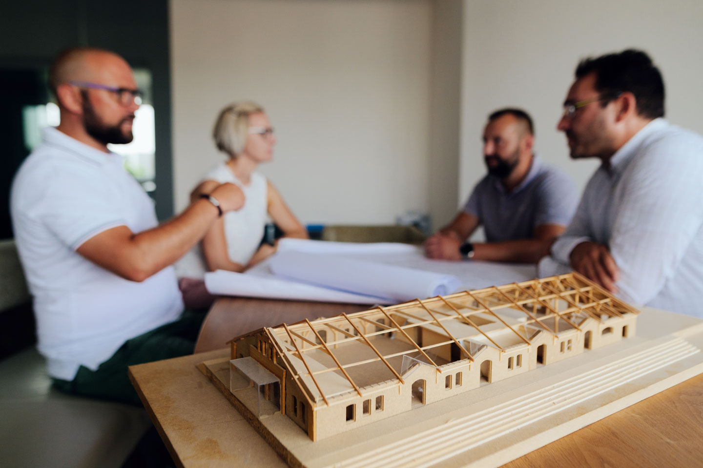 Ein Holzmodel eines großen Hauses steht im Vordergrund. Vier Personen sitzen an einem Besprechungstisch im Hintergrund.