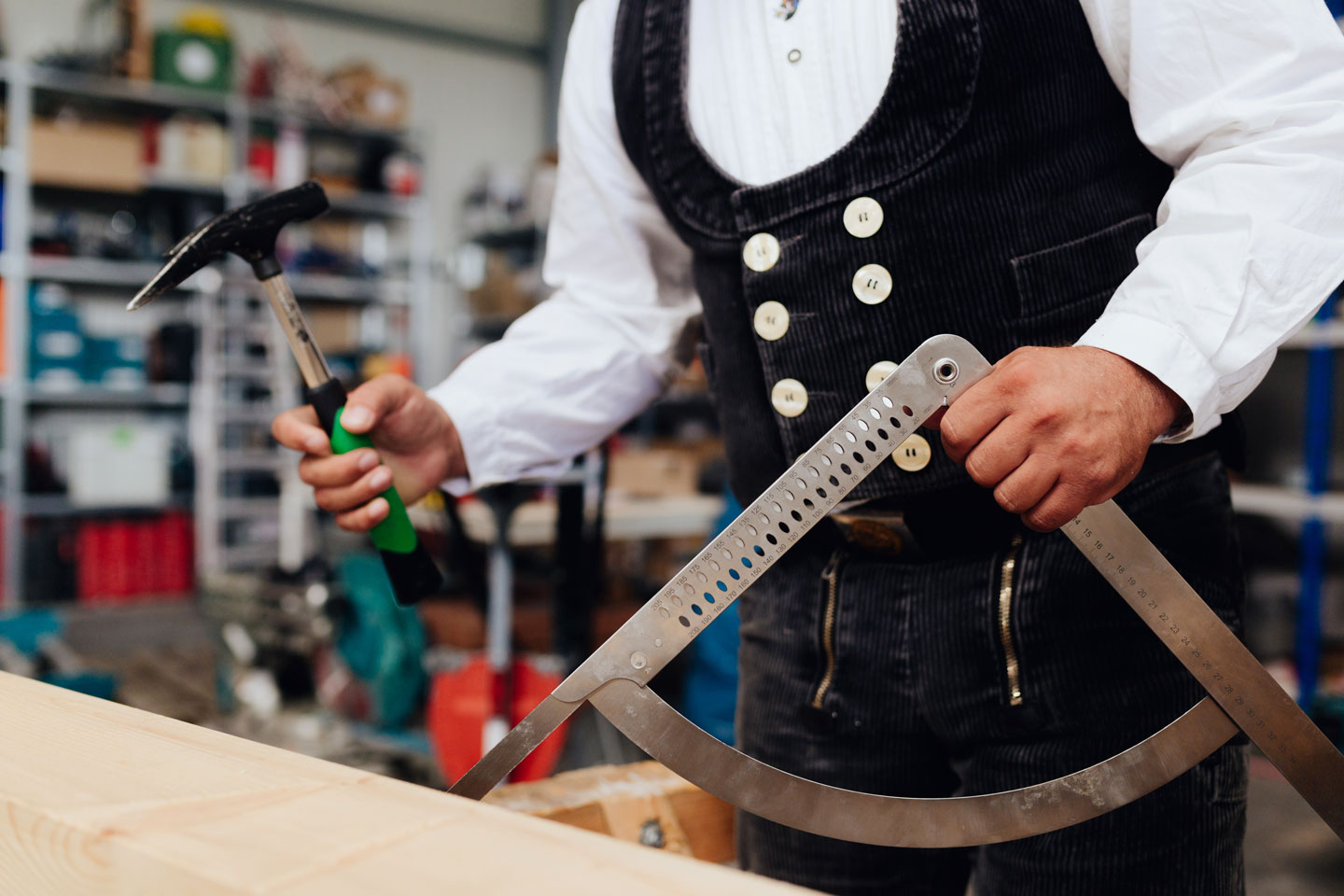 Zwei Werkzeuge des Zimmerers sind je in einer Hand einer Person in Zimmermannstracht – Winkelmesser und Hammer.