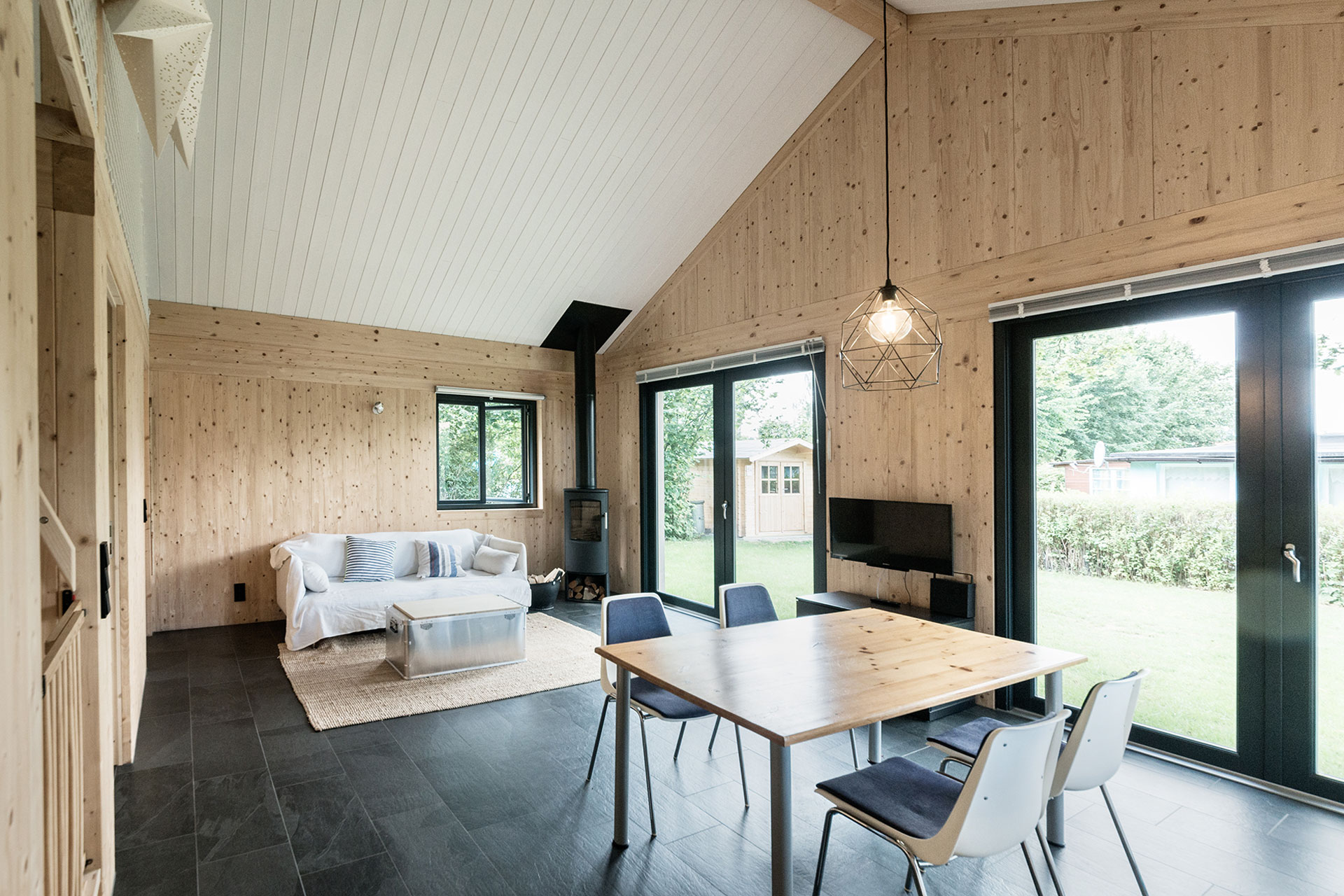 In einem Innenraum mit großen Fenstern stehen ein Holztisch, vier Stühle und ein Sofa.