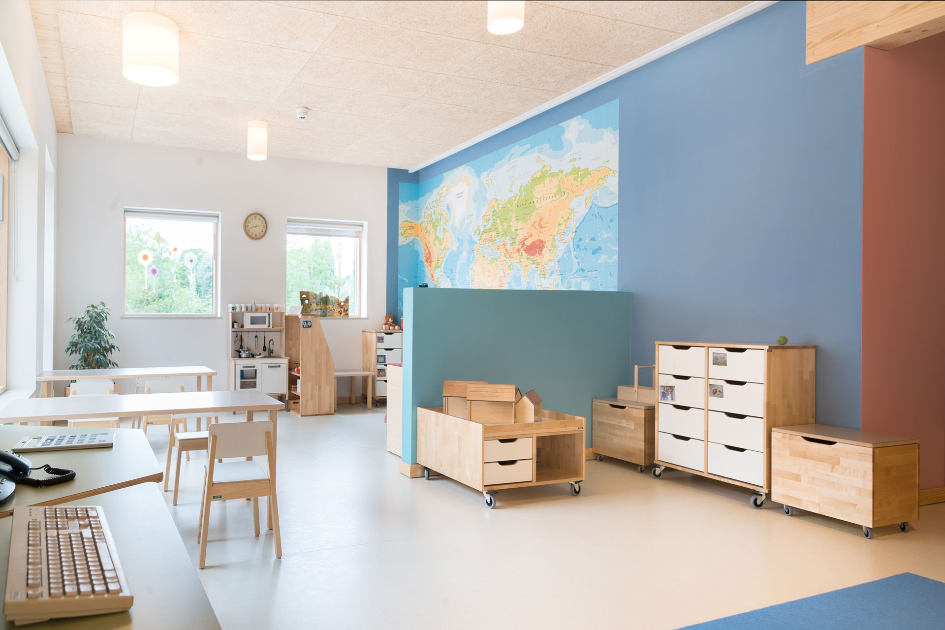 Ein heller Innenraum mit Spieleküche, Kinderstühlen und -tischen hat an einer blauen Wand eine sehr große Weltkarte.