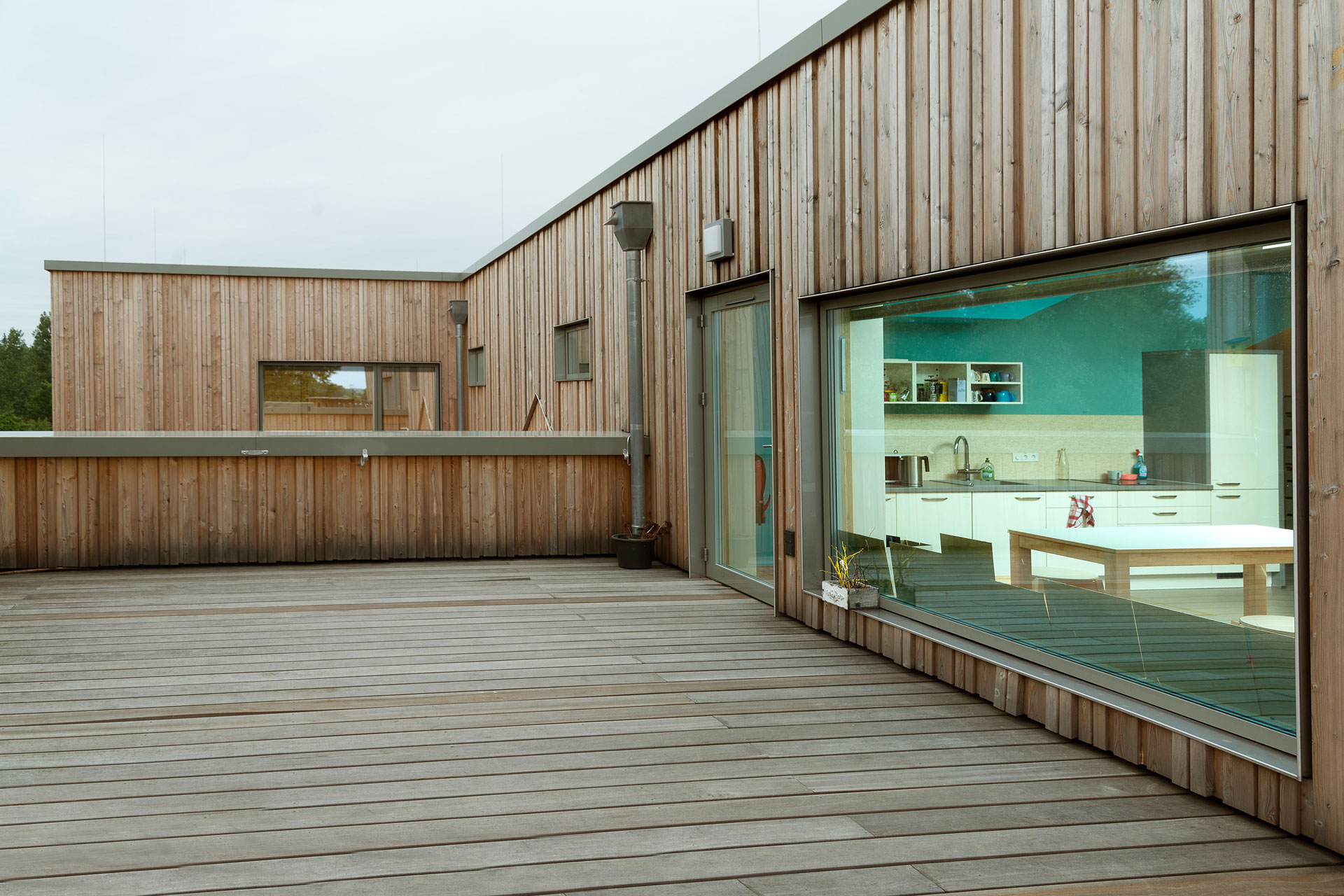 Von einer Dachterrasse, auch vollständig aus Holz, blickt man durch ein großes Fenster in einen Küchenbereich.