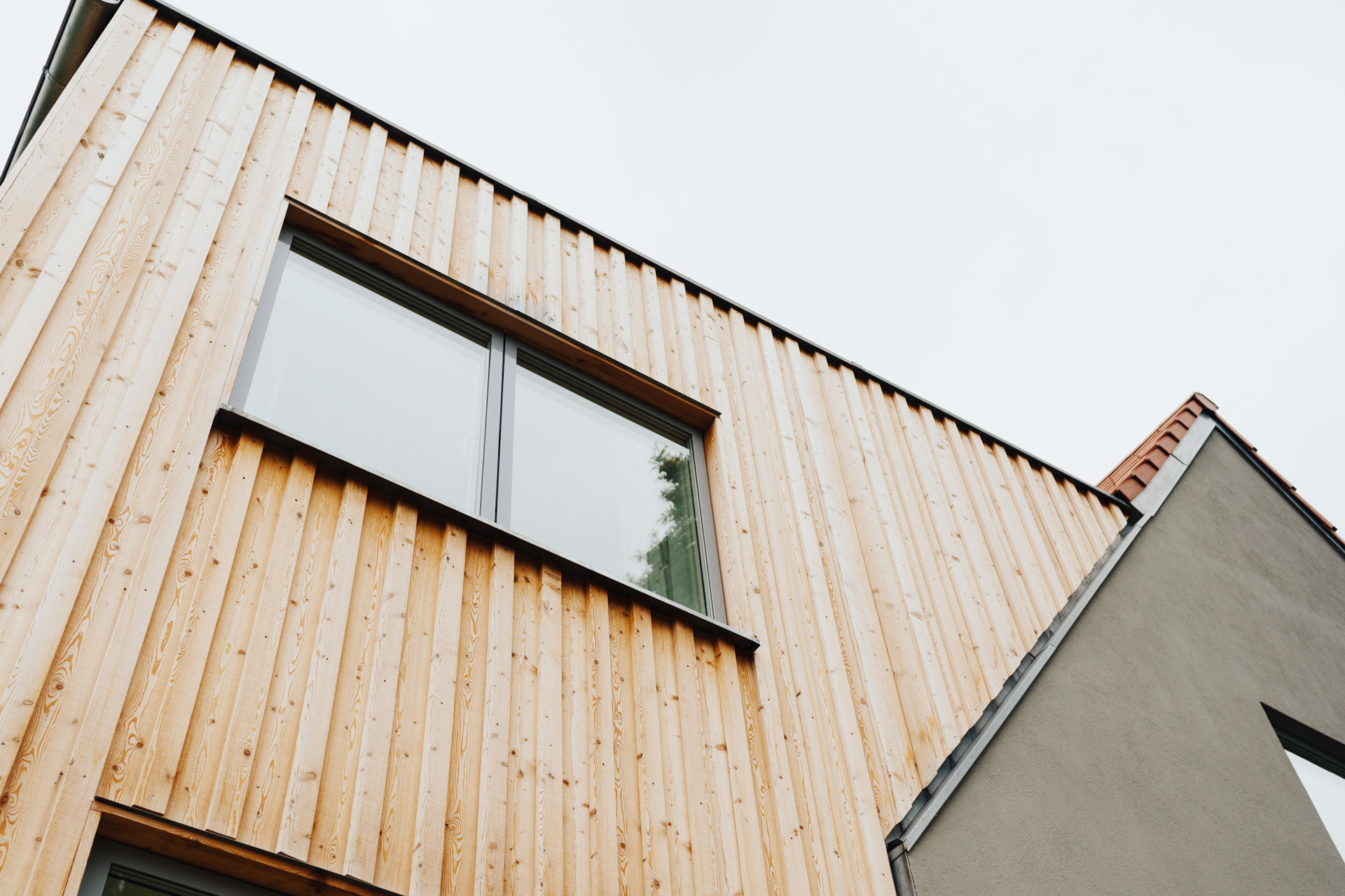 Die grau verputzte Fassade des Einfamilienhauses kontrastiert zum hellen Holz des Anbaus.