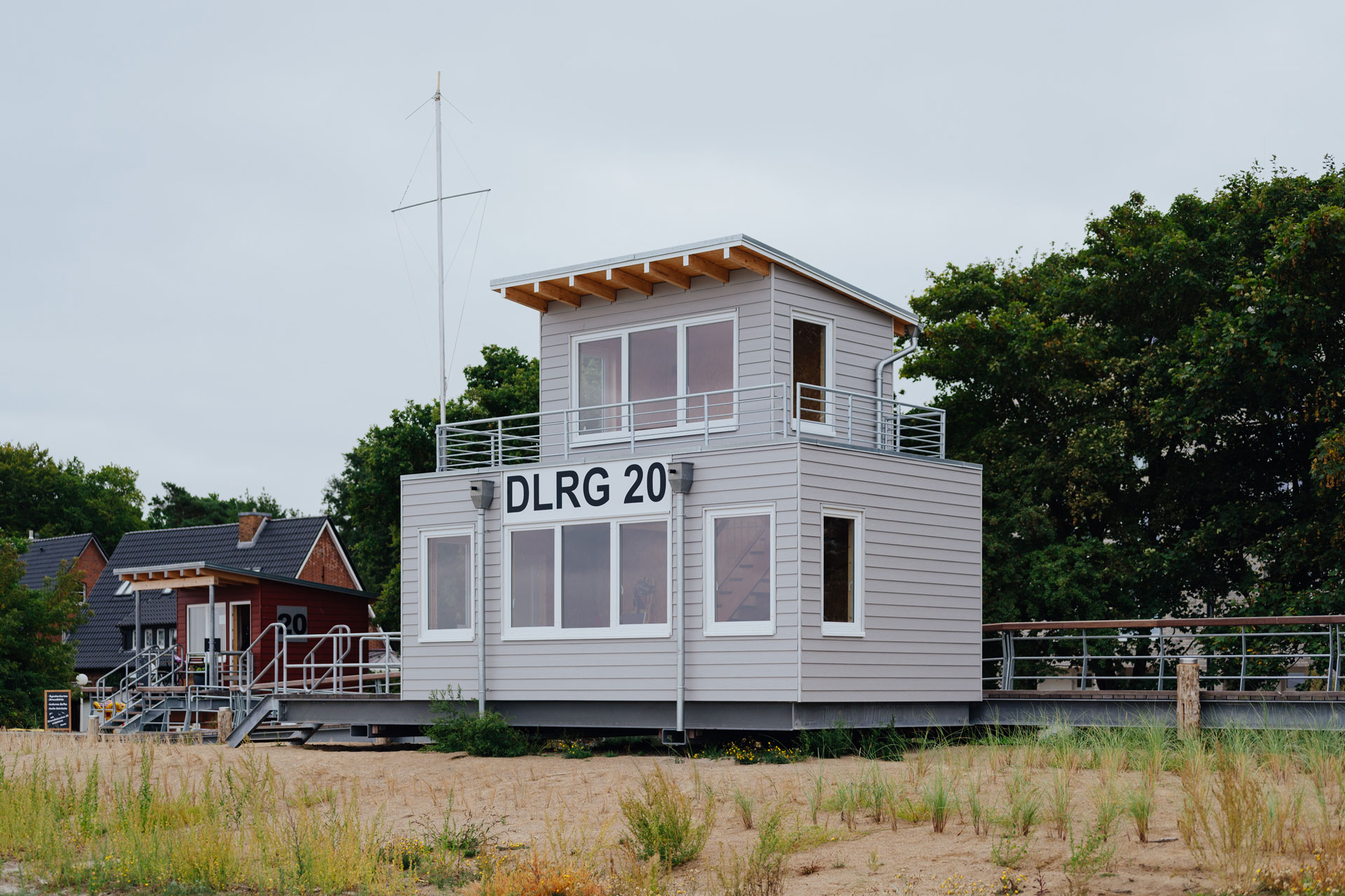 An einem Strandhaus mit hellgrau gestrichenen Holz steht die Nummer DLRG 20 auf einem Schild.