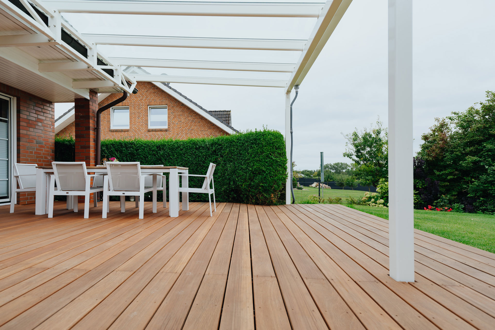 Auf einer großen Holzterrasse steht ein Tisch mit sechs Stühlen. Ein grüne Hecke und Rasen umgibt die Terrasse.