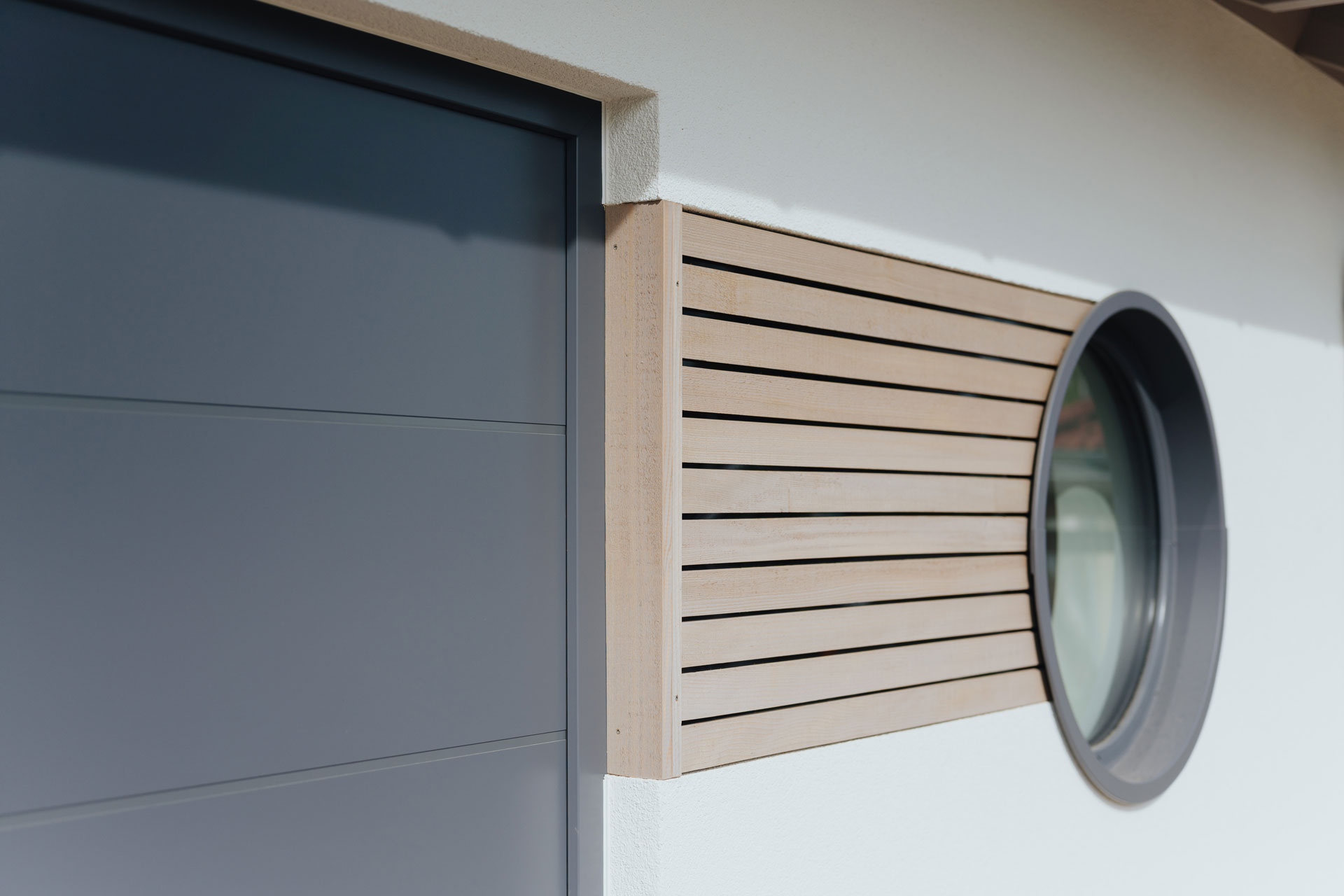 Ein kleines rundes Fenster hat eine seitliche Holzverkleidung auf einer sonst weißen Fassade.
