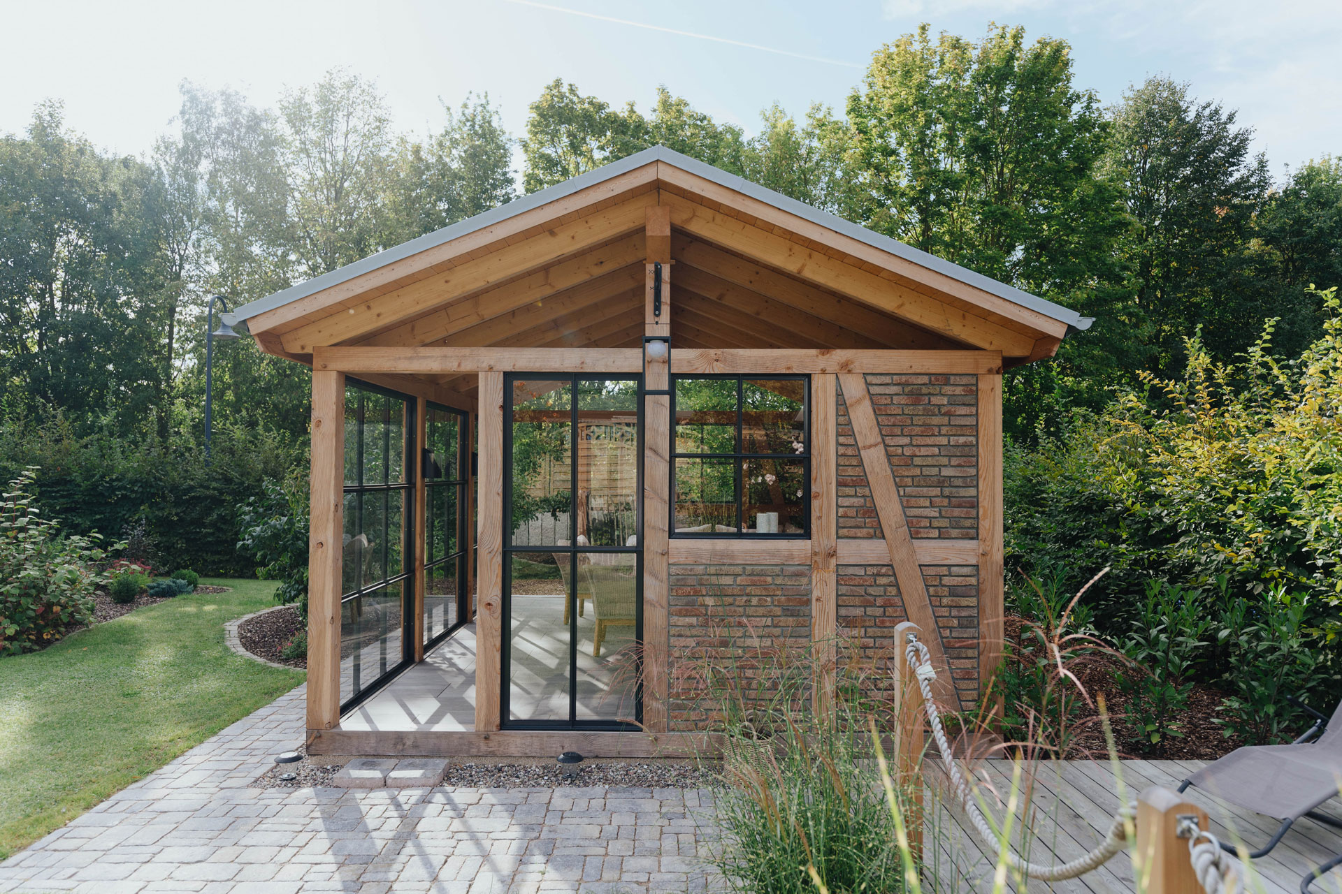 Das Gartenhaus mit dominanten Holzbalken, etwas Mauerwerk und viel Glas ist umgeben von viel Grün.
