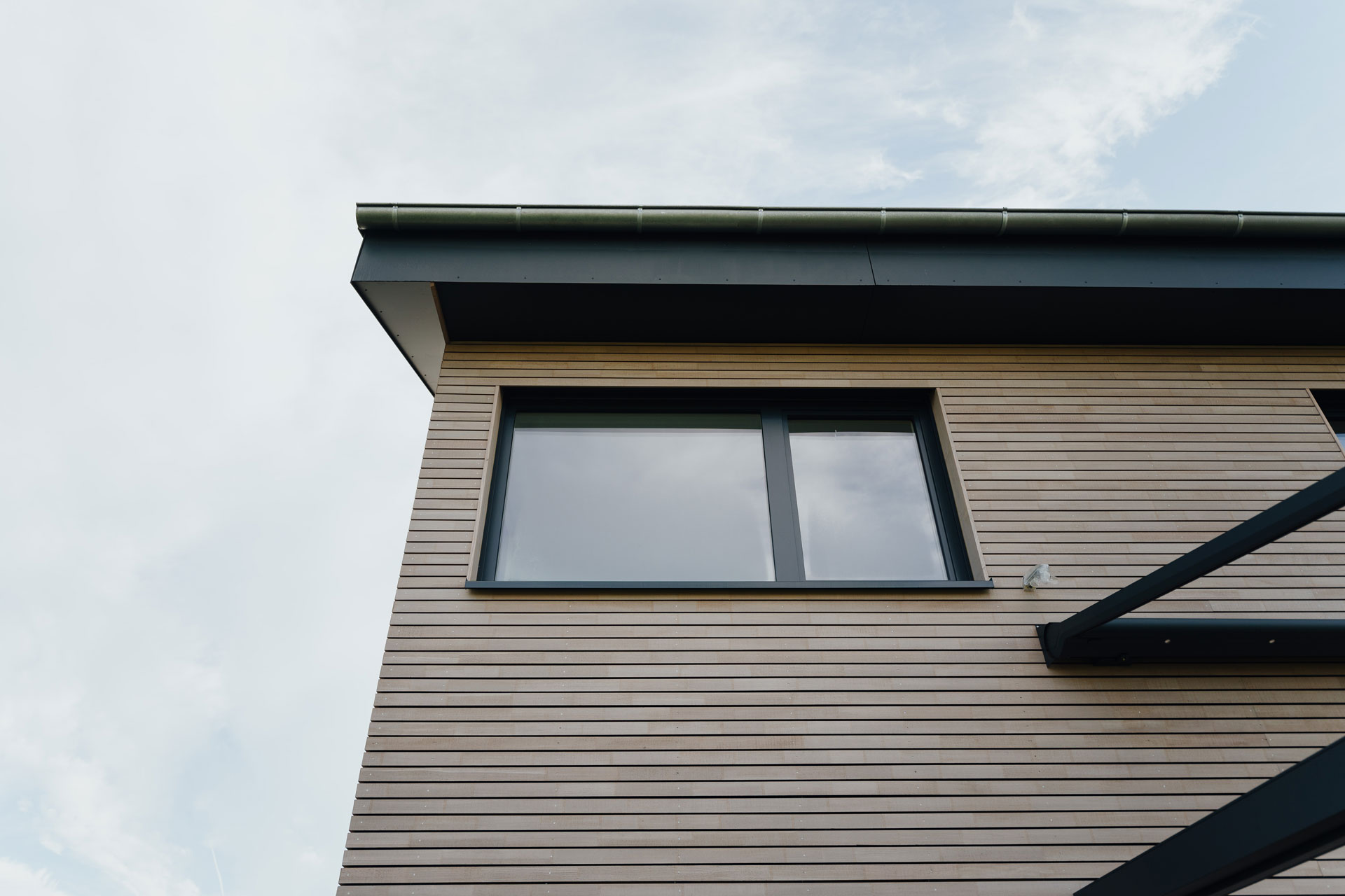 Verkleidete Holzfassade und angrenzendes Dach sind vor leicht bewölkten Himmel aufgenommen.