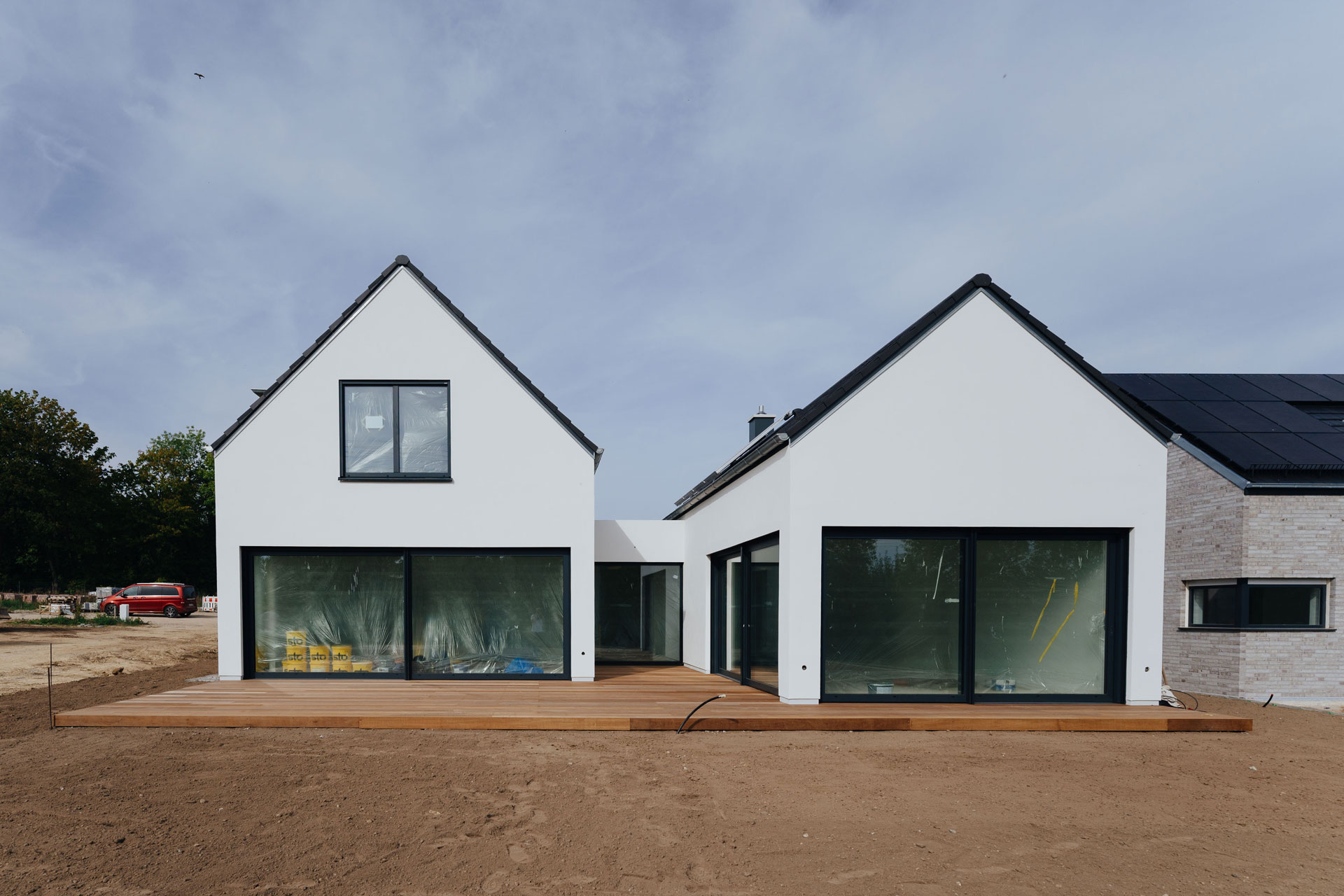 Ein Einfamilienhaus hat einen großzügige Holzterrasse. Zwei Hausbauten sind mit einem Übergang mit großen Glasflächen verbunden.